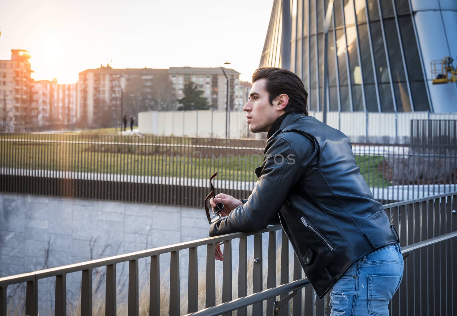 One handsome young man in urban setting in modern city, standing, wearing black leather jacket and jeans