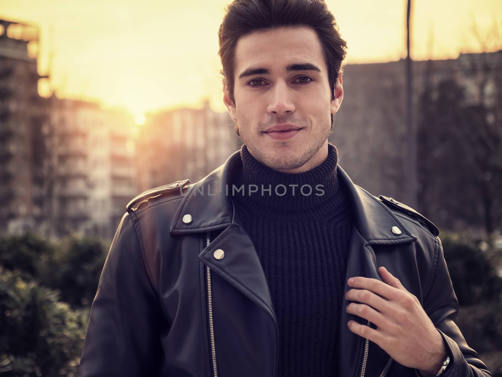 One handsome young man in urban setting in modern city, standing, wearing black leather jacket and jeans, looking at camera, at sunset