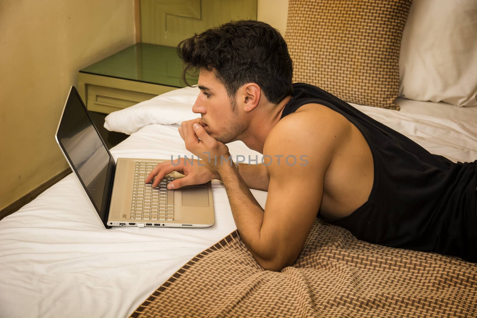 Attractive Young Man with Serious Expression, Reclining Comfortably with Laptop on Bed Working on his Start-up Business - Young Male College or University Student Doing Homework, in Bedroom