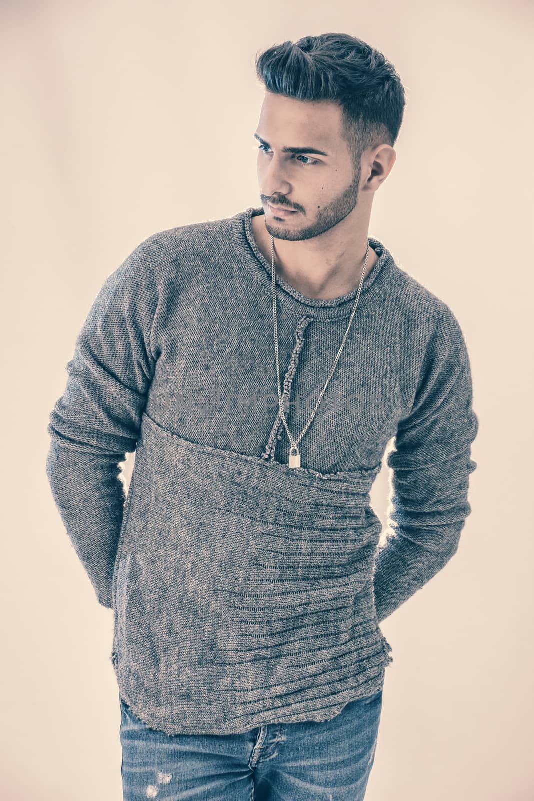 Handsome young man wearing grey sweater and ripped jeans, on light background in studio shot