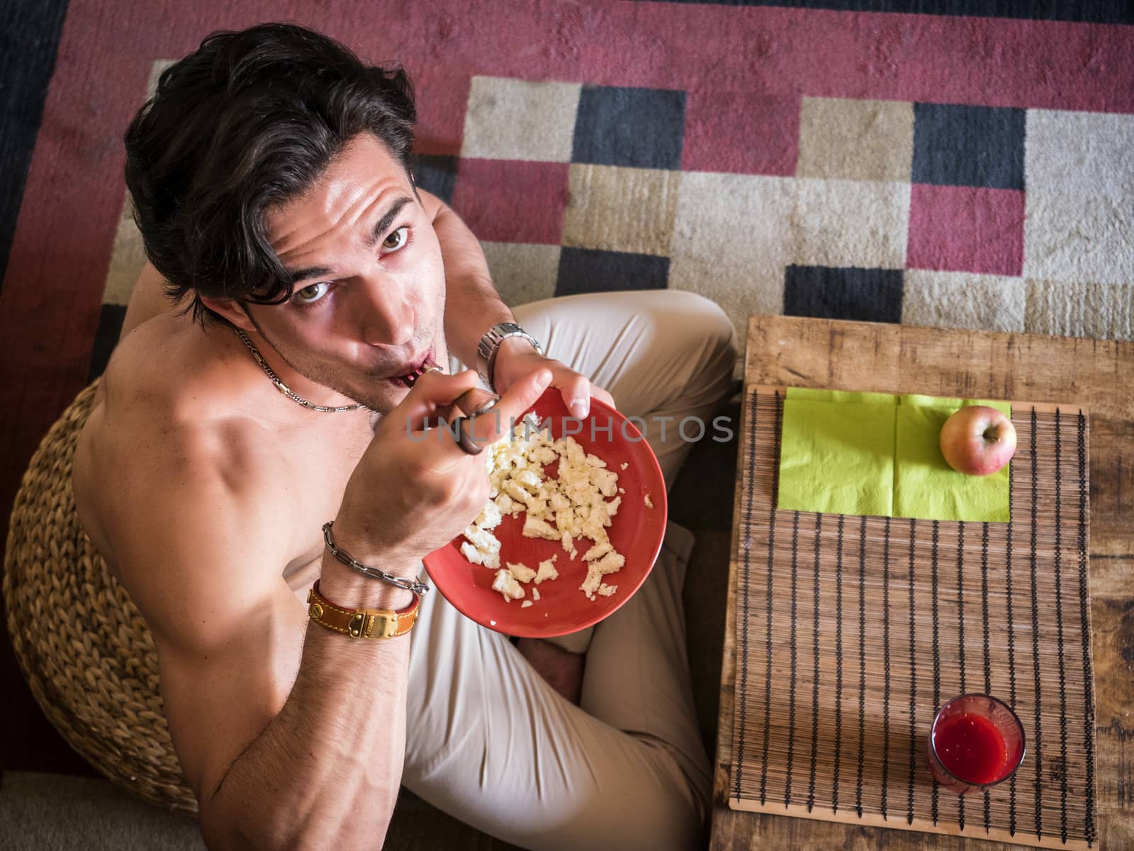 From above shot of happy muscular shirtless man sitting on floor having breakfast eating cereal, smiling at camera