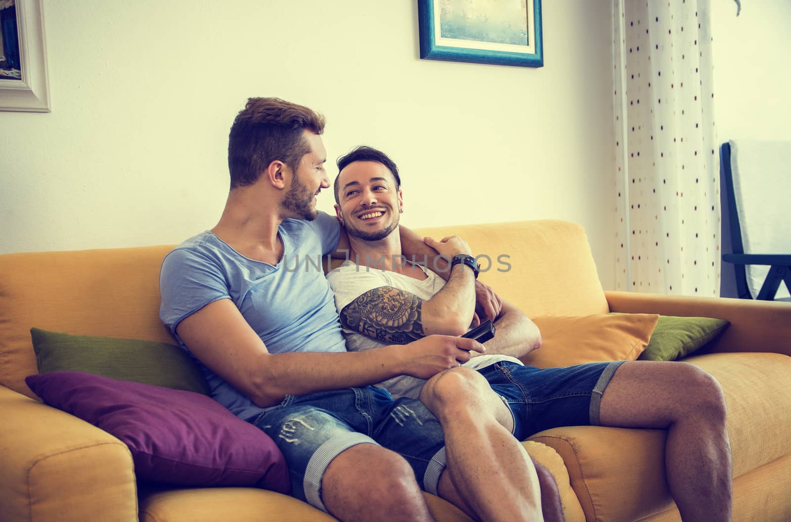 Two gay men on sofa embracing at home by artofphoto