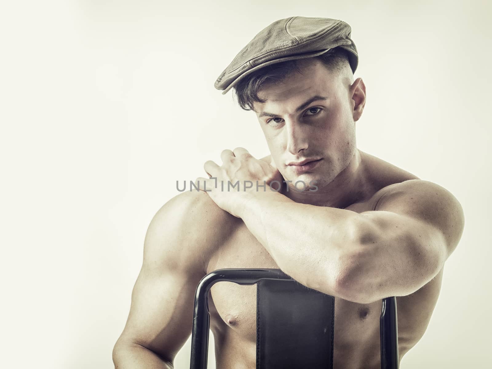 Muscular bodybuilder sitting on chair, looking at camera, wearing hat