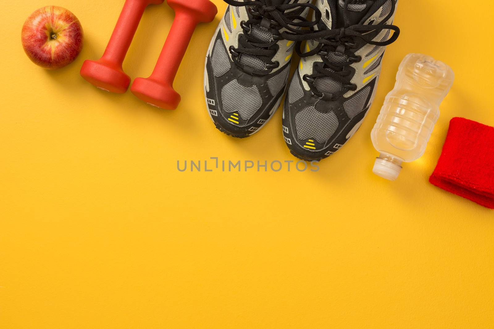Sport shoes, dumbbells, apple, water bottle and wrist bands. Fitness still life, on yellow background with copy space.