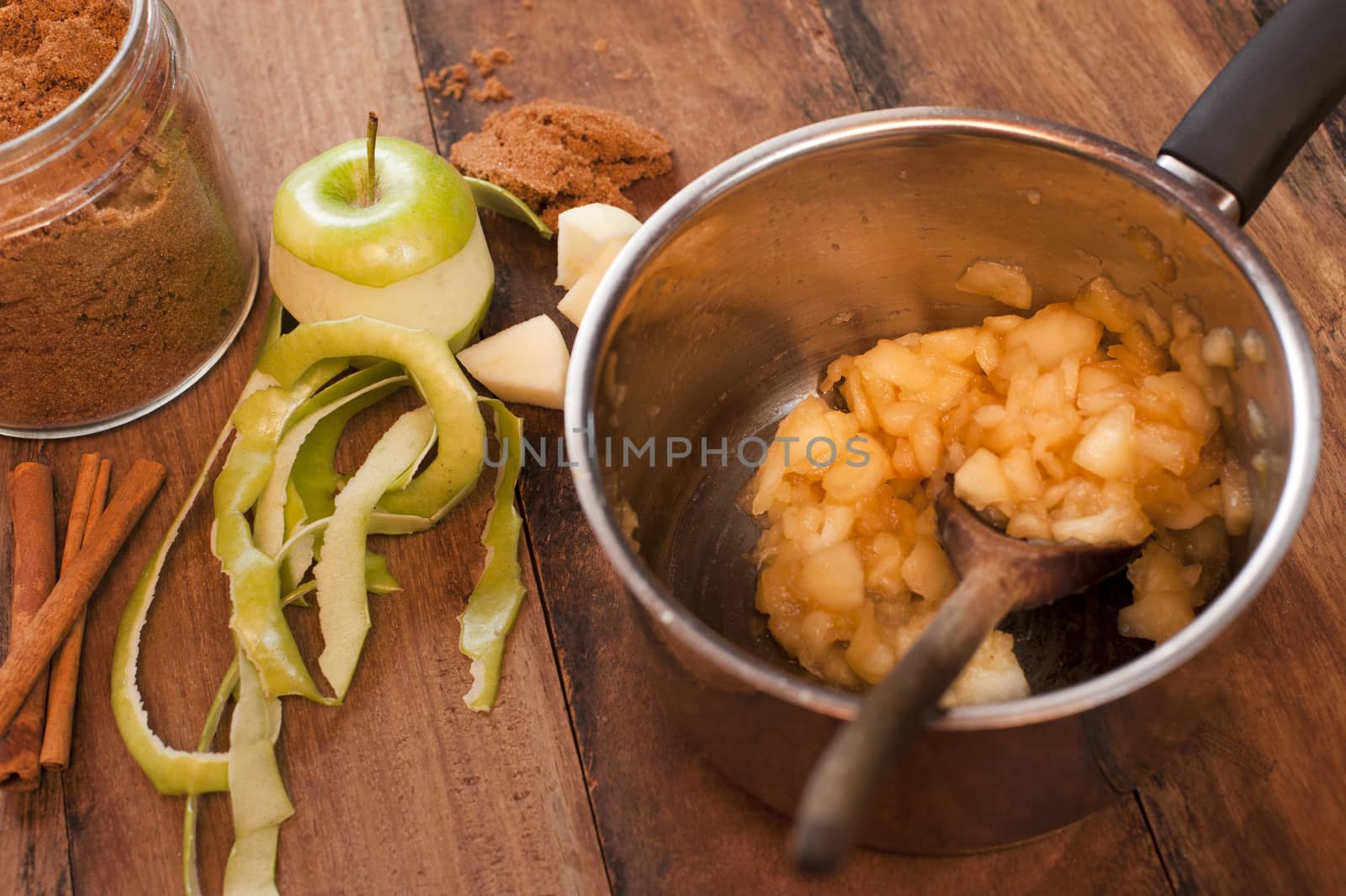 Pieces of apple in metal pan with wooden spoon and apple peeled on table, next to jar of cinnamon, cooking homemade apple sauce