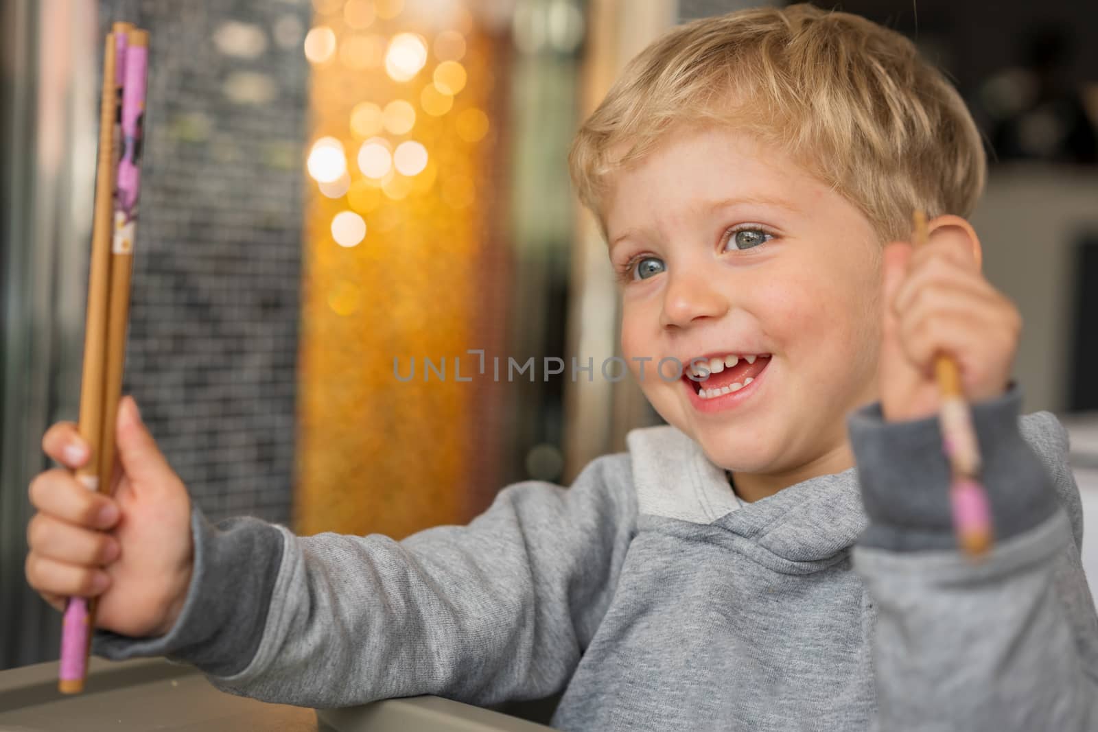 Baby boy sitting in high chair plays with chopsticks and smiling happiness at chinese restaurant.Natural light background with nice bokeh,horizontal photo.