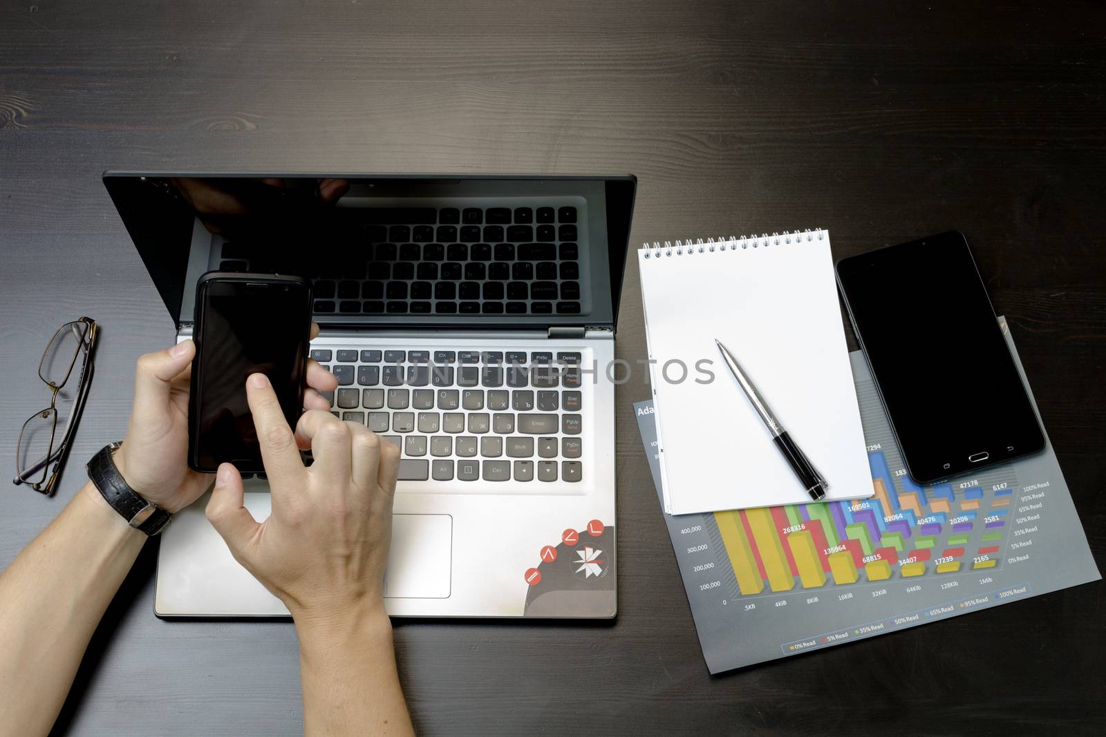 Businessman working on an ultra-thin laptop. Nearby are a block, a marker, a pen, glasses, a tablet phone. On his hand is an expensive watch.