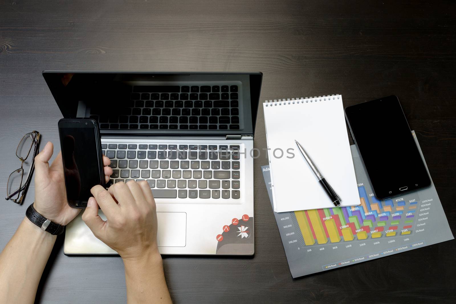 Businessman working on an ultra-thin laptop. Nearby are a block, a marker, a pen, glasses, a tablet phone. On his hand is an expensive watch.