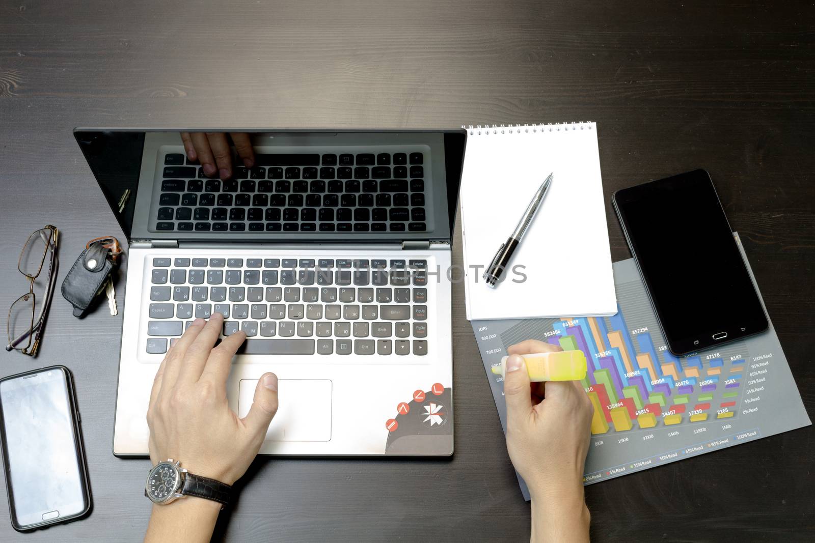 Businessman working on an ultra-thin laptop. Nearby are a block, a marker, a pen, glasses, a tablet phone. On his hand is an expensive watch.