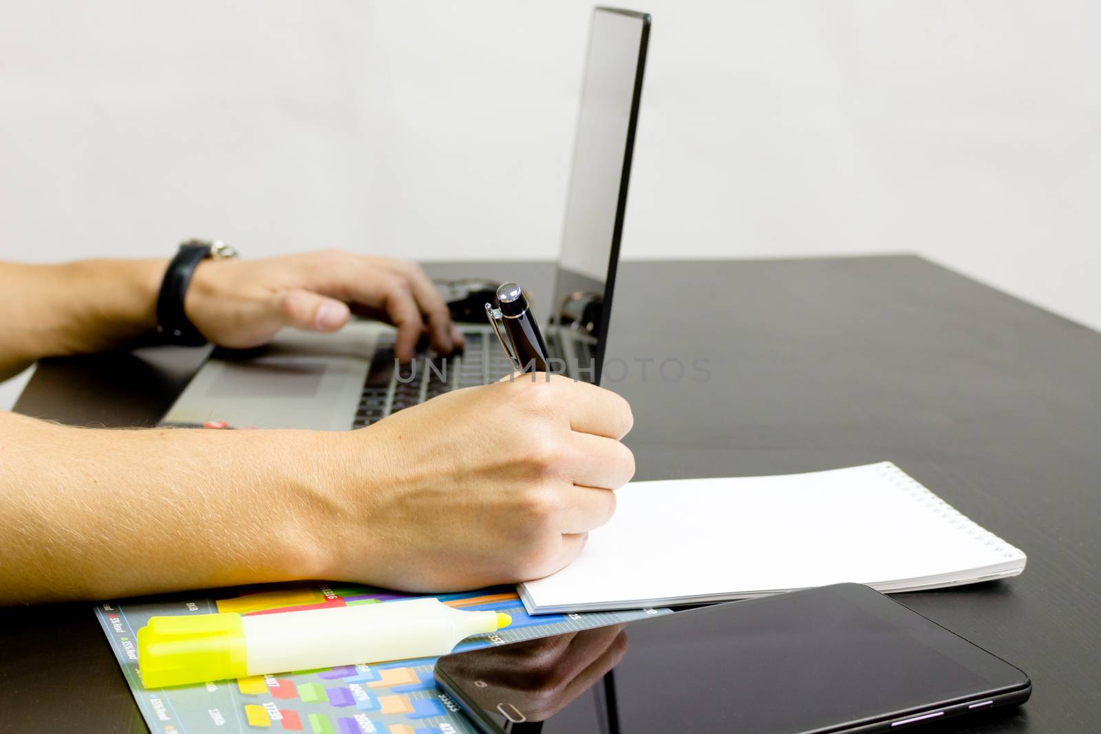 Businessman working on an ultra-thin laptop. Nearby are a block, a marker, a pen, glasses, a tablet phone. On his hand is an expensive watch.