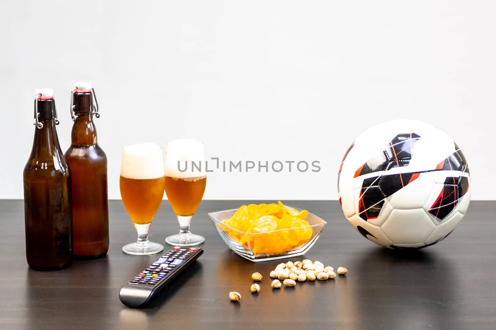 People prepared to watch football on TV with beer. There's beer on the table, ball, TV remote, snacks. Craft beer. Light background.