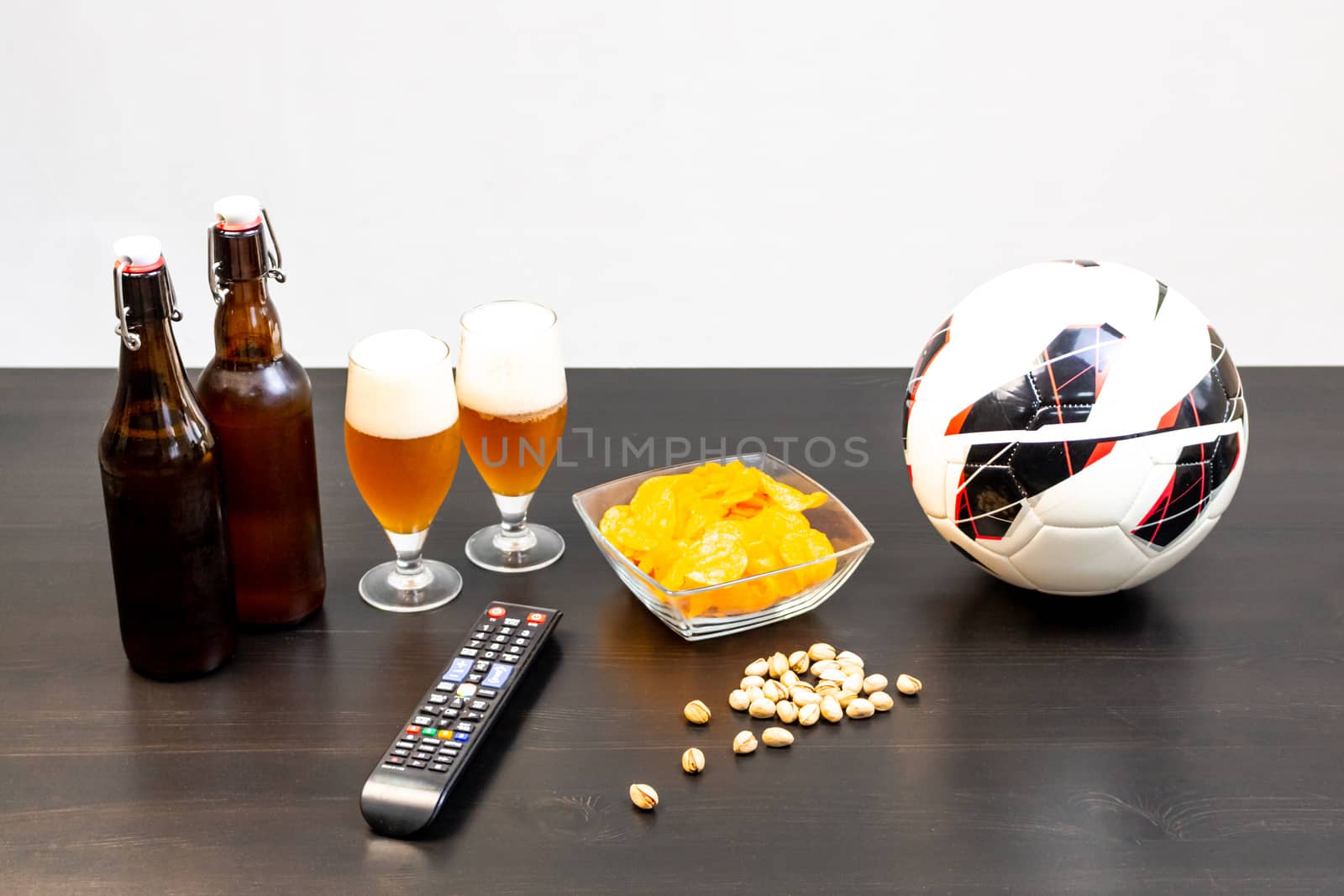 People prepared to watch football on TV with beer. There's beer on the table, ball, TV remote, snacks. Craft beer. Light background.