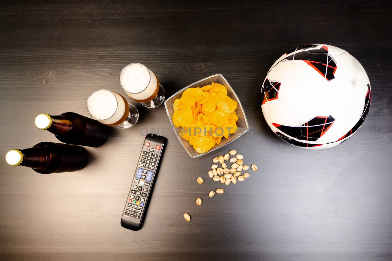 People prepared to watch football on TV with beer. There's beer on the table, ball, TV remote, snacks. Craft beer. Light background. The view from the top.