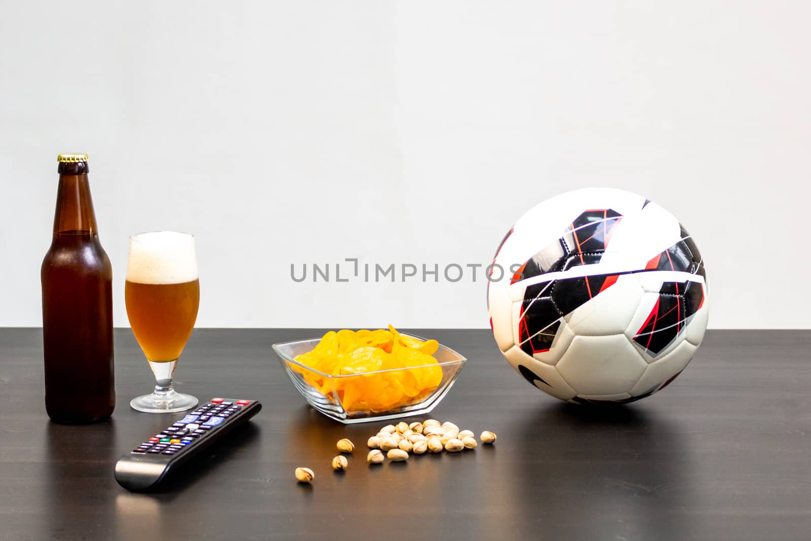 People prepared to watch football on TV with beer. There's beer on the table, ball, TV remote, snacks. Craft beer. Light background.