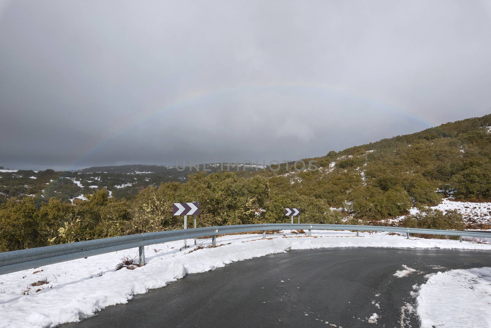 Mountain road on a cloudy day on winter season, rainbow in the background. by HERRAEZ