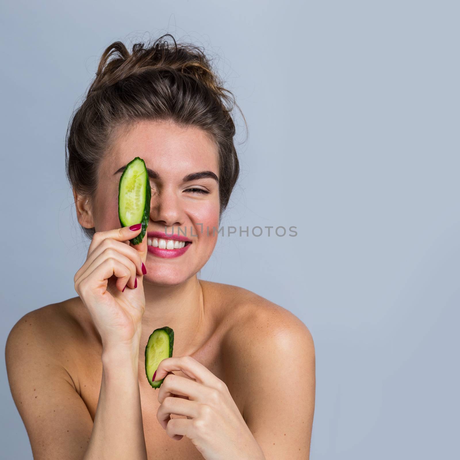 Smiling woman holding slices of cucumber in front of her eyes