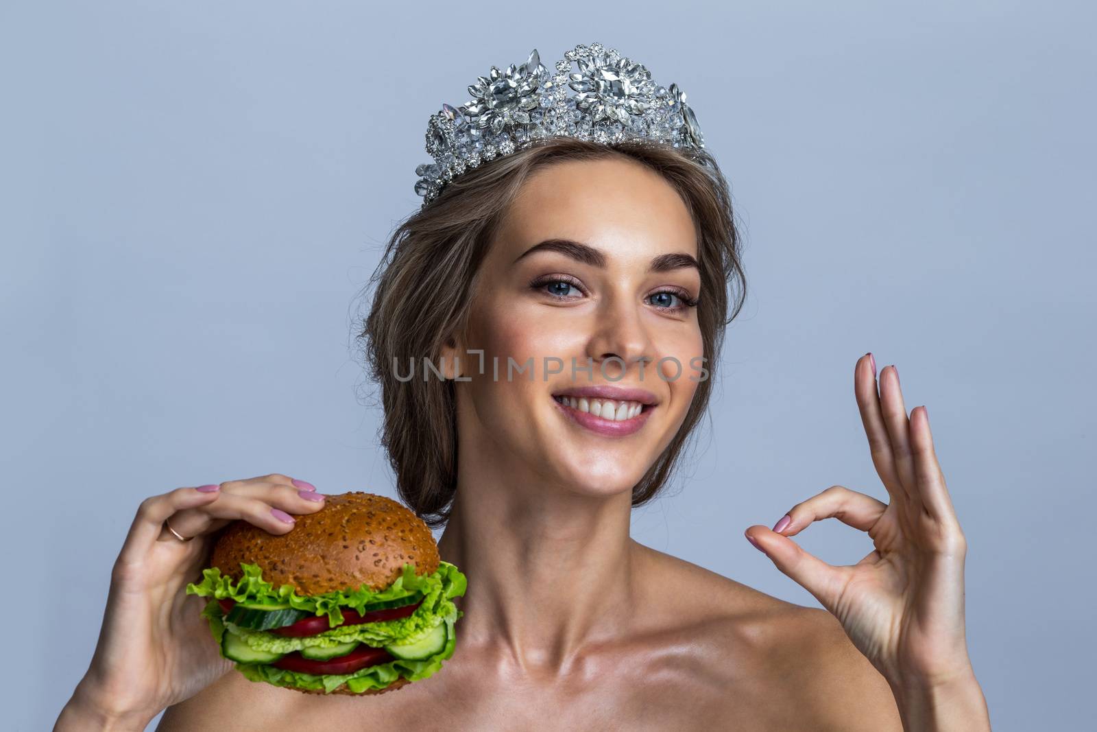 Portrait of beautiful young blonde woman with a diadem on a head holding vegan burger made of vegetables and showing ok sign