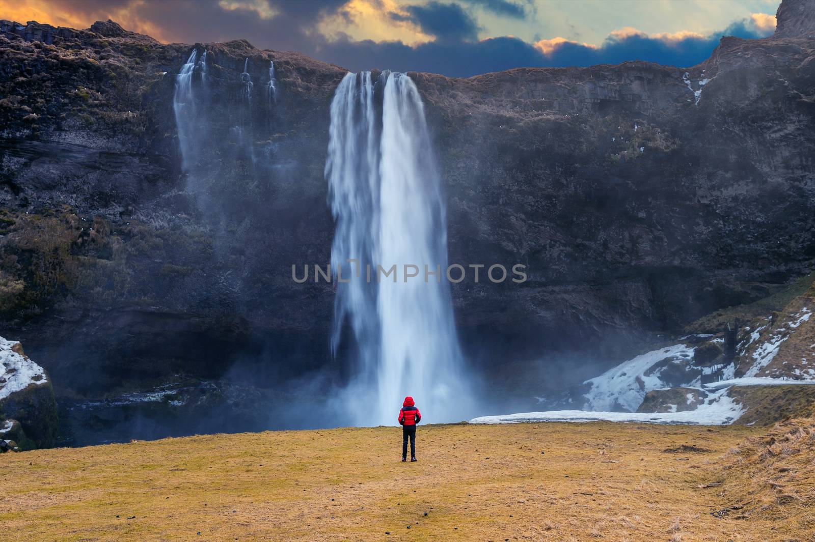 Seljalandsfoss waterfall in Iceland. Guy in red jacket looks at Seljalandsfoss waterfall. by gutarphotoghaphy