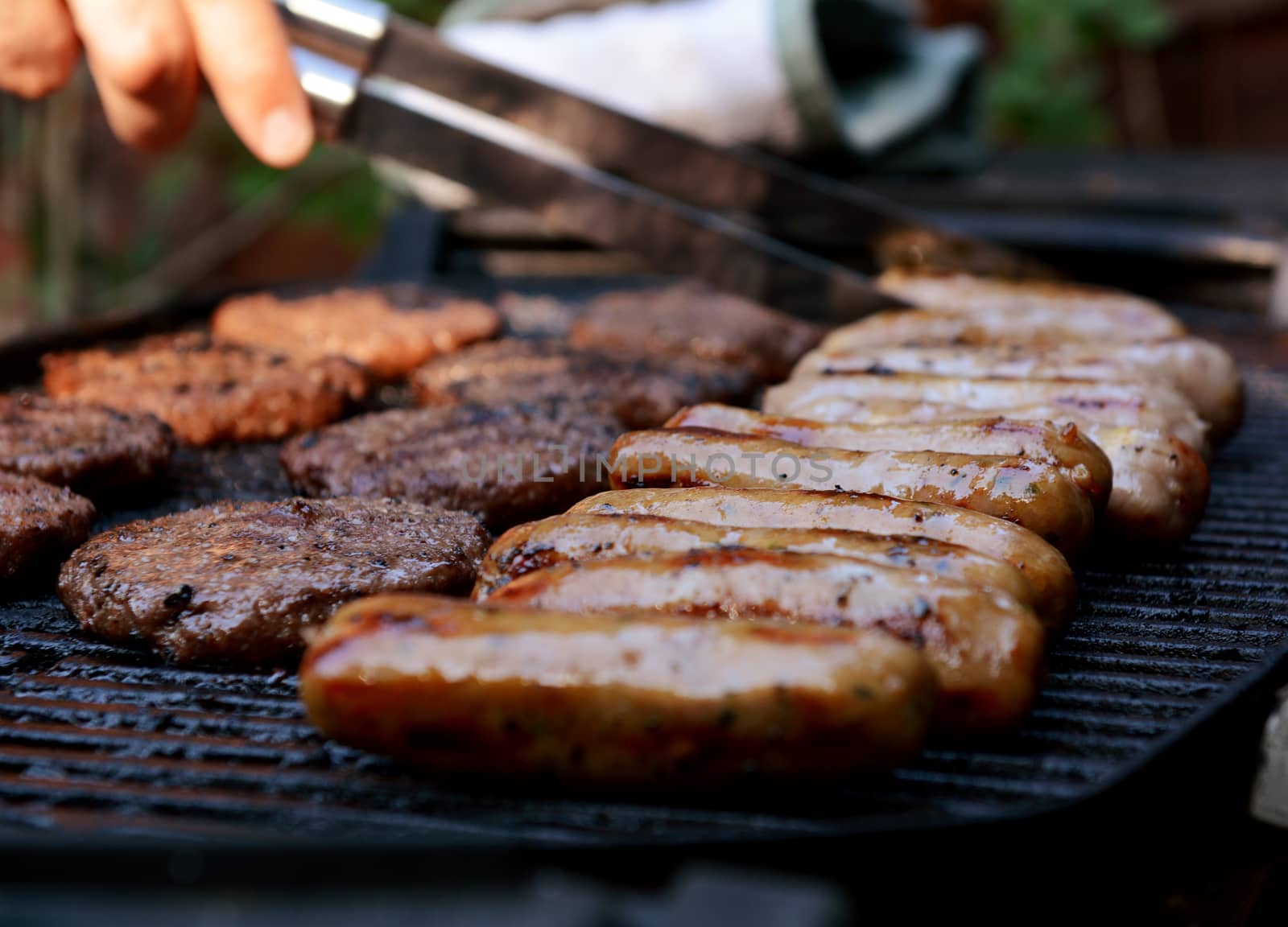 Sausages and hamburgers on a barbecue grill, the cook uses tongs in the background 