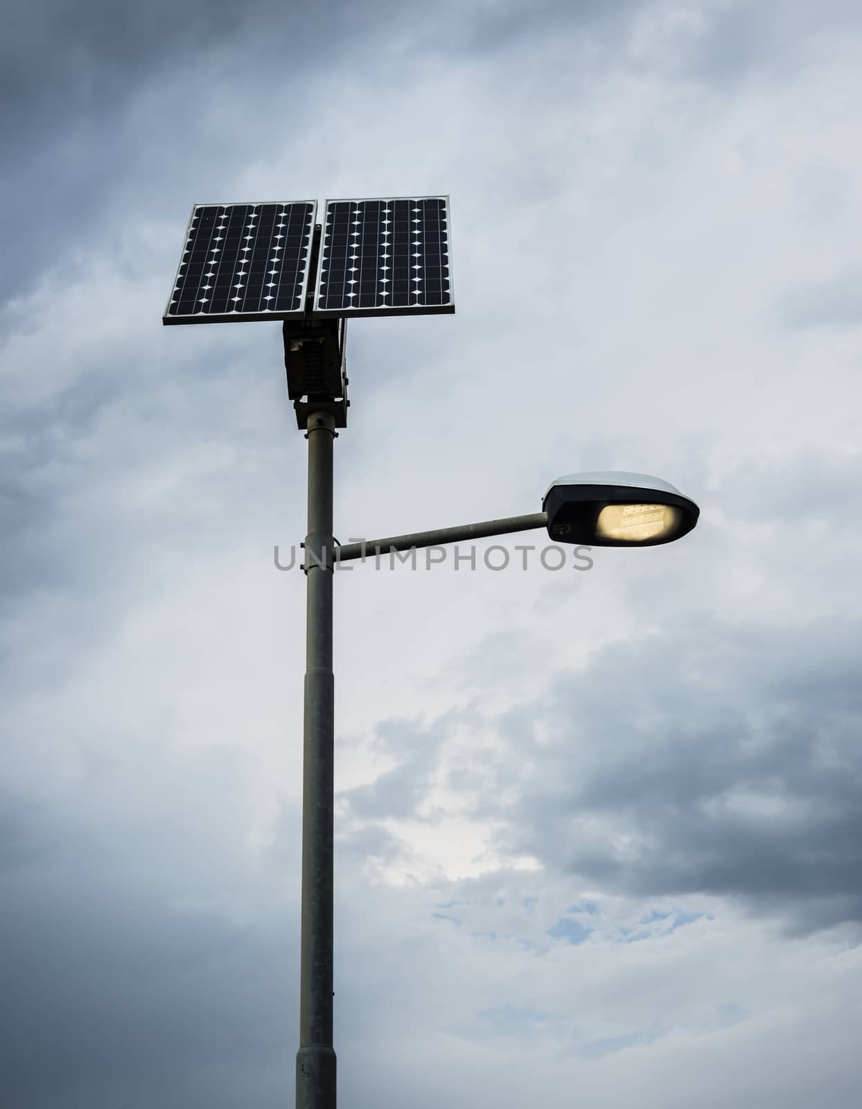 Solar panel on street lamp post with light on and cloudy sky.