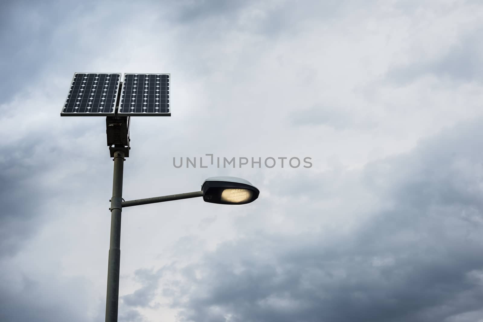 Solar panel on street lamp post with light on and cloudy sky.