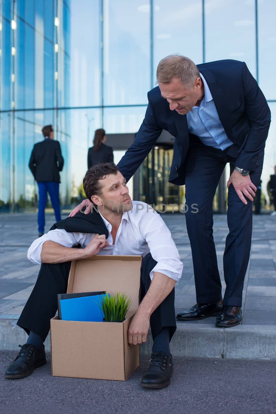 Fired business man sitting frustrated and upset on the street near office building with box of his belongings. He lost work. Other businessman comforts and encourages him