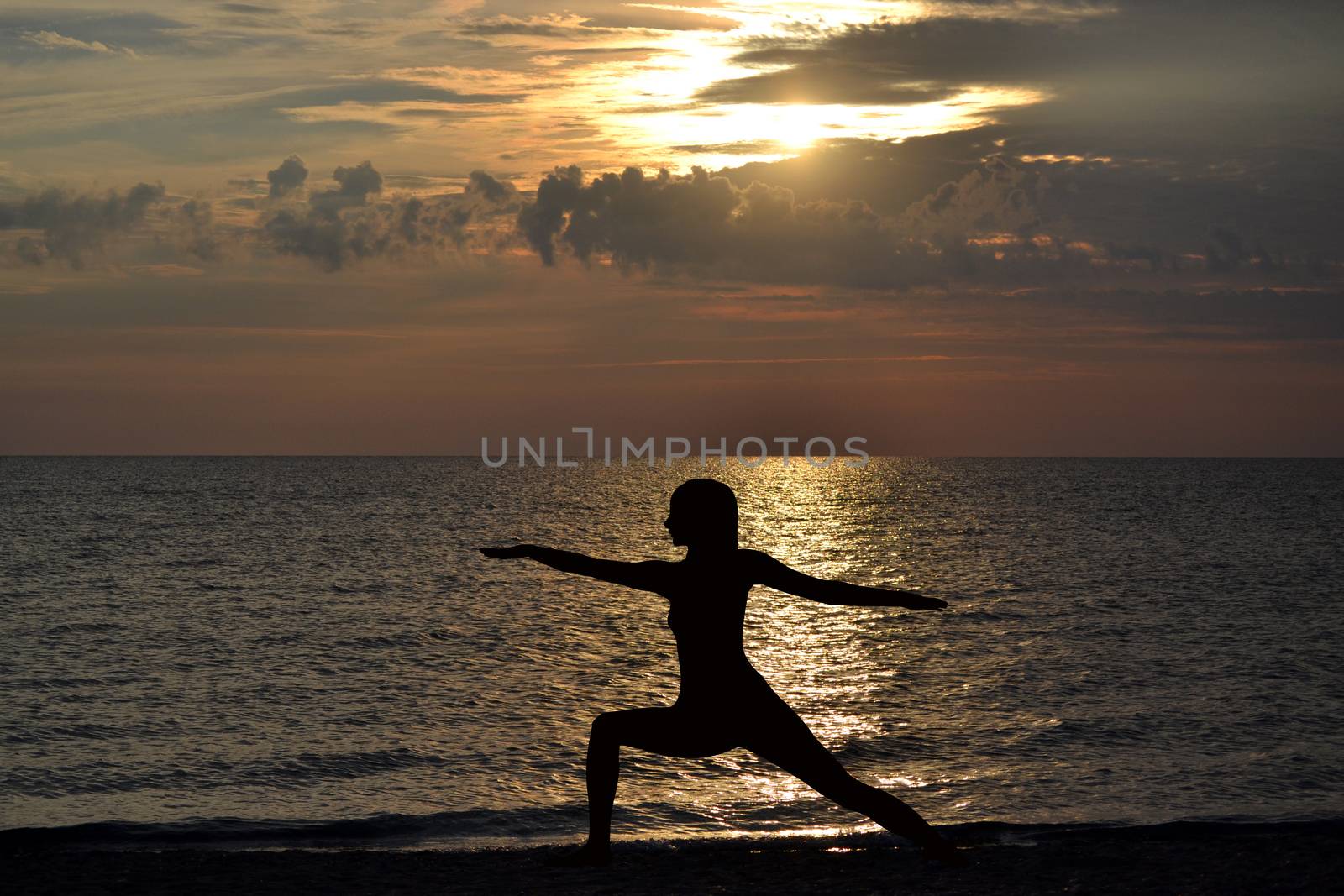 Woman practicing yoga, standing in Virabhadrasana pose at sunset