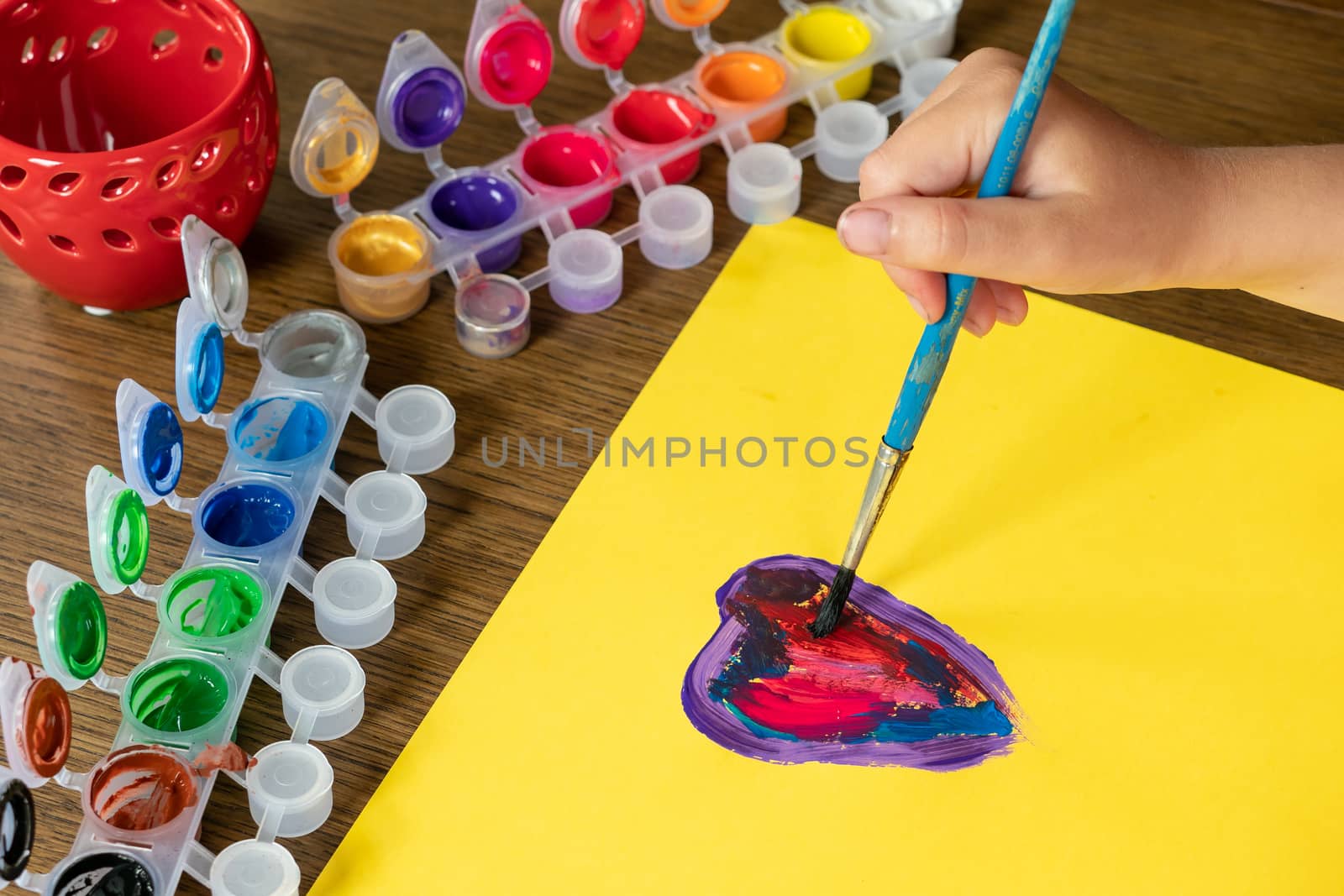 Child painting a picture. A little girl’s hand painting a colorful heart on yellow paper.