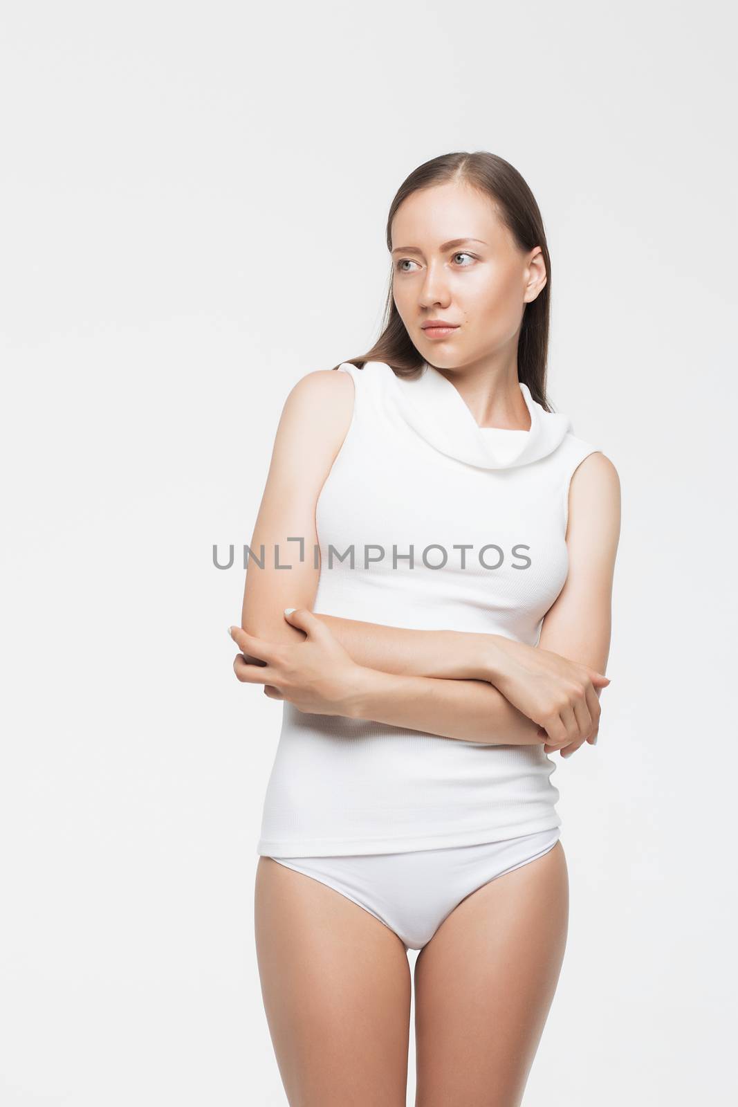 Beautiful young woman in panties and turtleneck top keeping arms folded and looking away while standing on white background