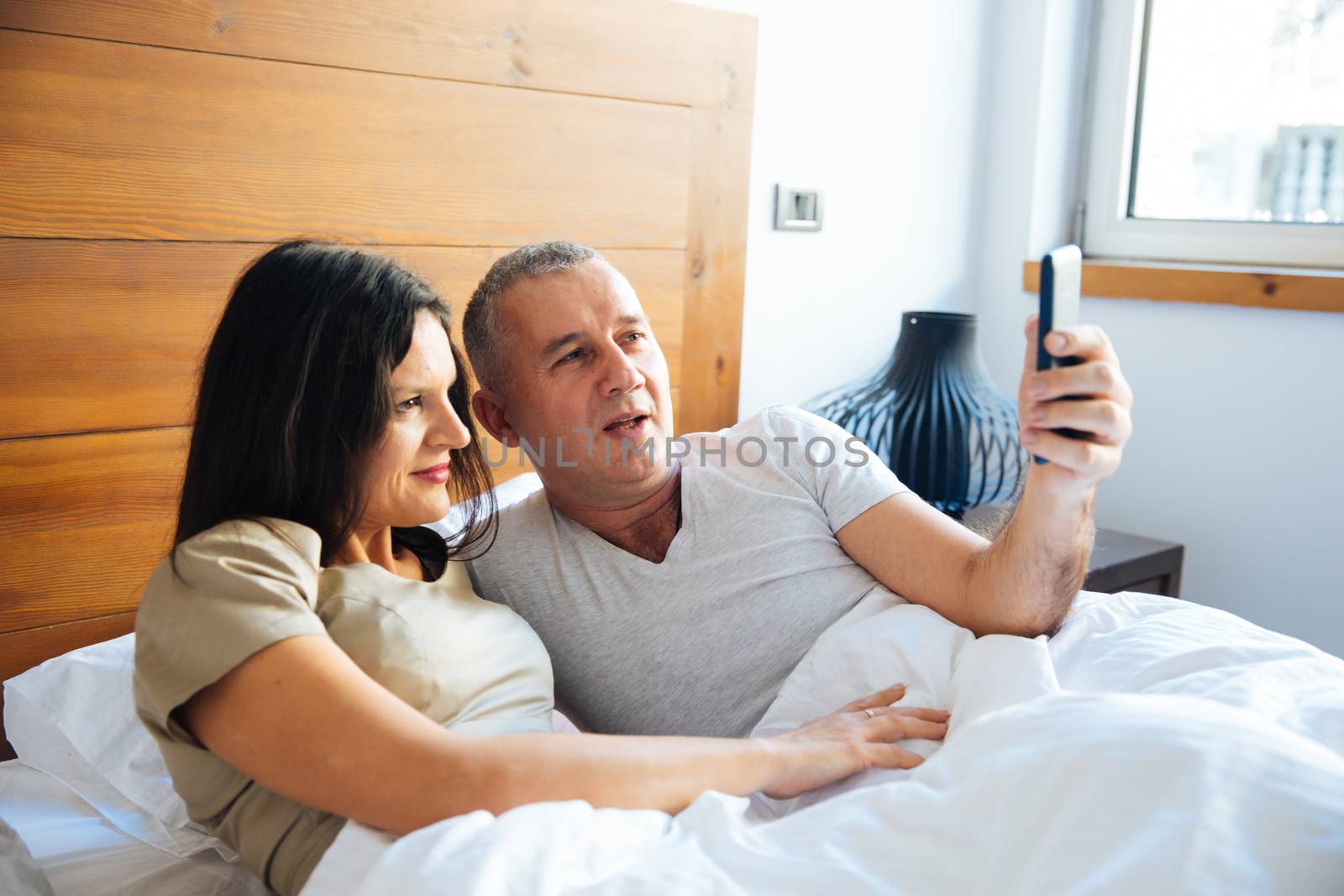 Attractive and Happy Mid Aged Couple Enjoying in the Bed in the Morning and Using a Smartphone and Taking Selfie Photo