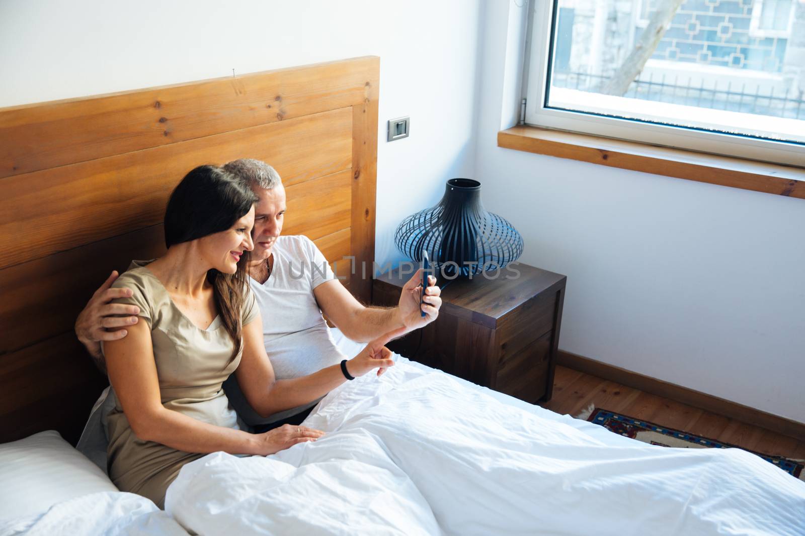 Attractive and Happy Mid Aged Couple Enjoying in the Bed in the Morning and Using a Smartphone
