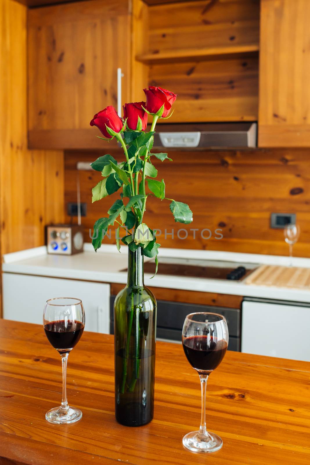 Three Red Roses In Bottle And Two Glasses Of Red Wine On Wooden Table