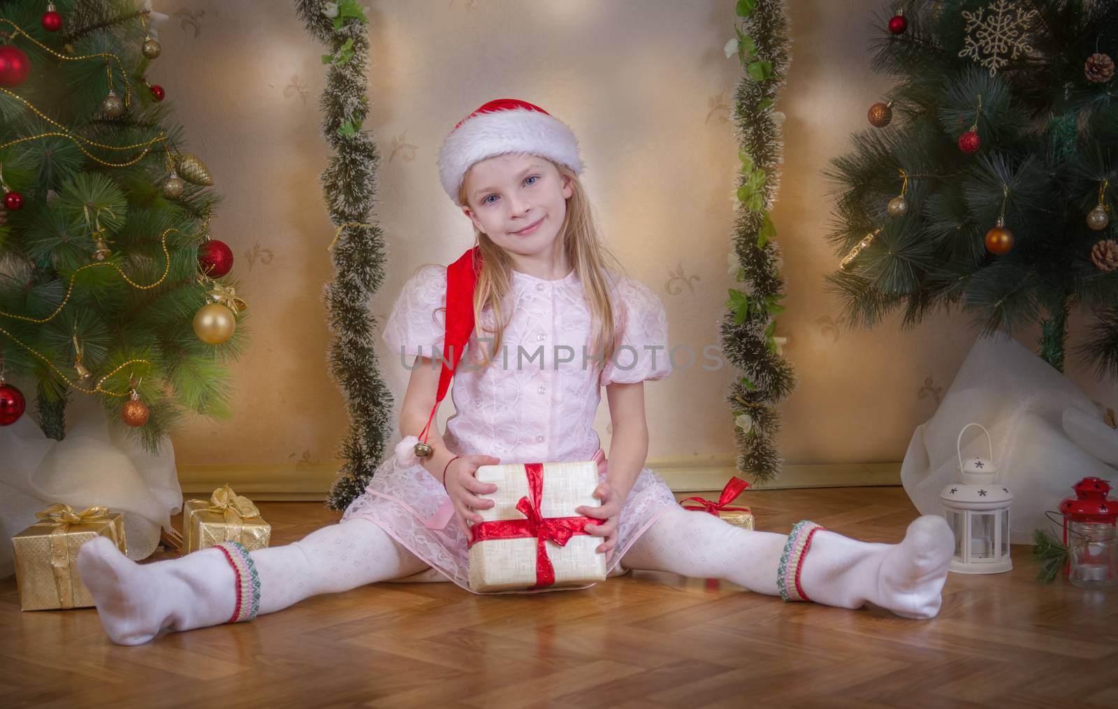 Cute girl in Santa hat sitting with gift among Christmas tree