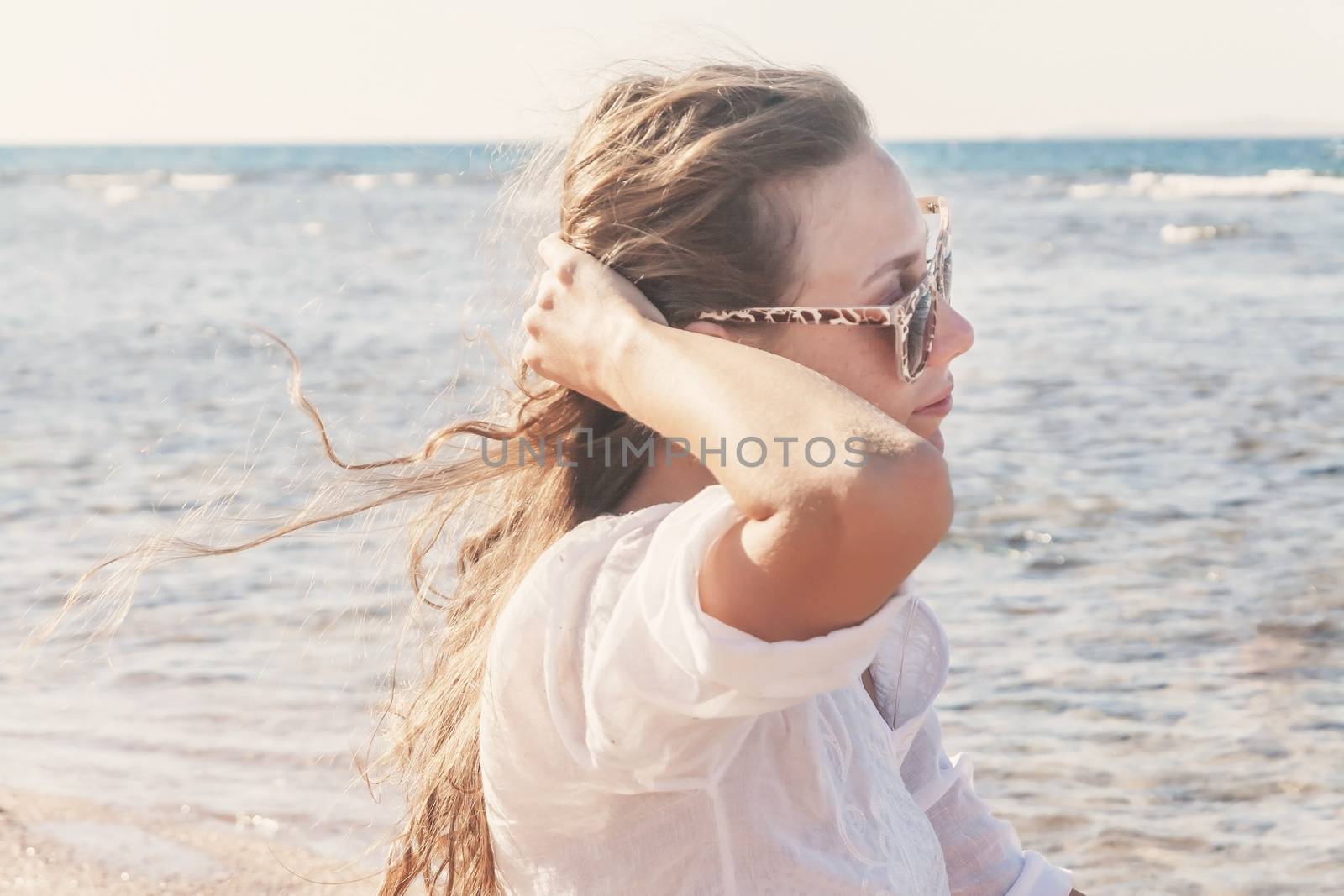 A young woman in sunglasses holds a hand to her hair fluttering in the wind near the sea, toning in retro style. The concept of a free way of life.