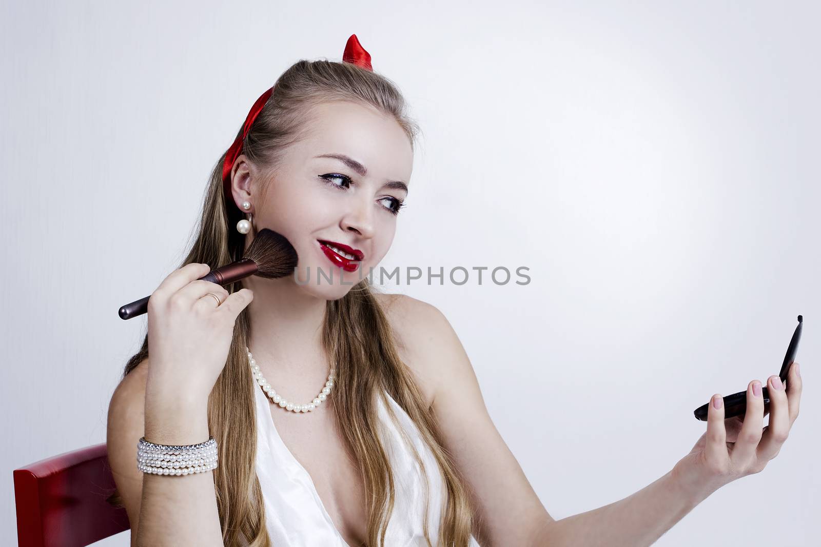 Young girl brushing her face with makeup while holding small mirror in other hand
