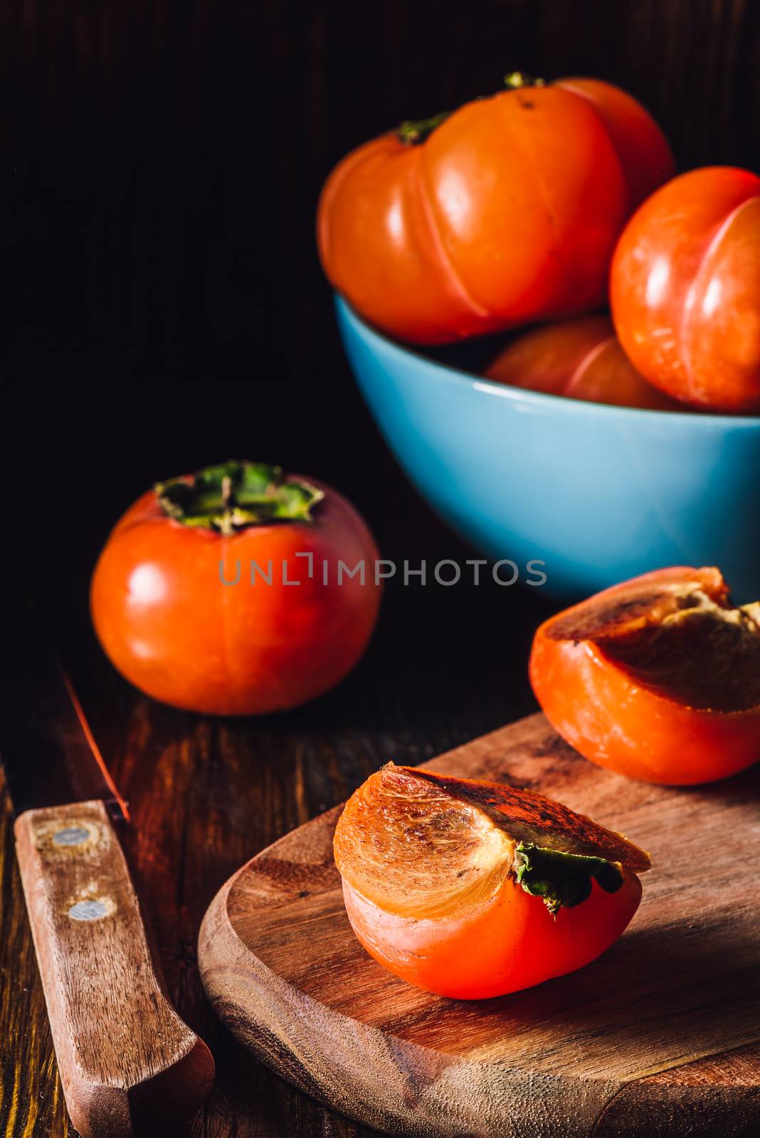 Persimmon Slices with Knife and Some Fruits in Blue Bowl on Background. Vertical.