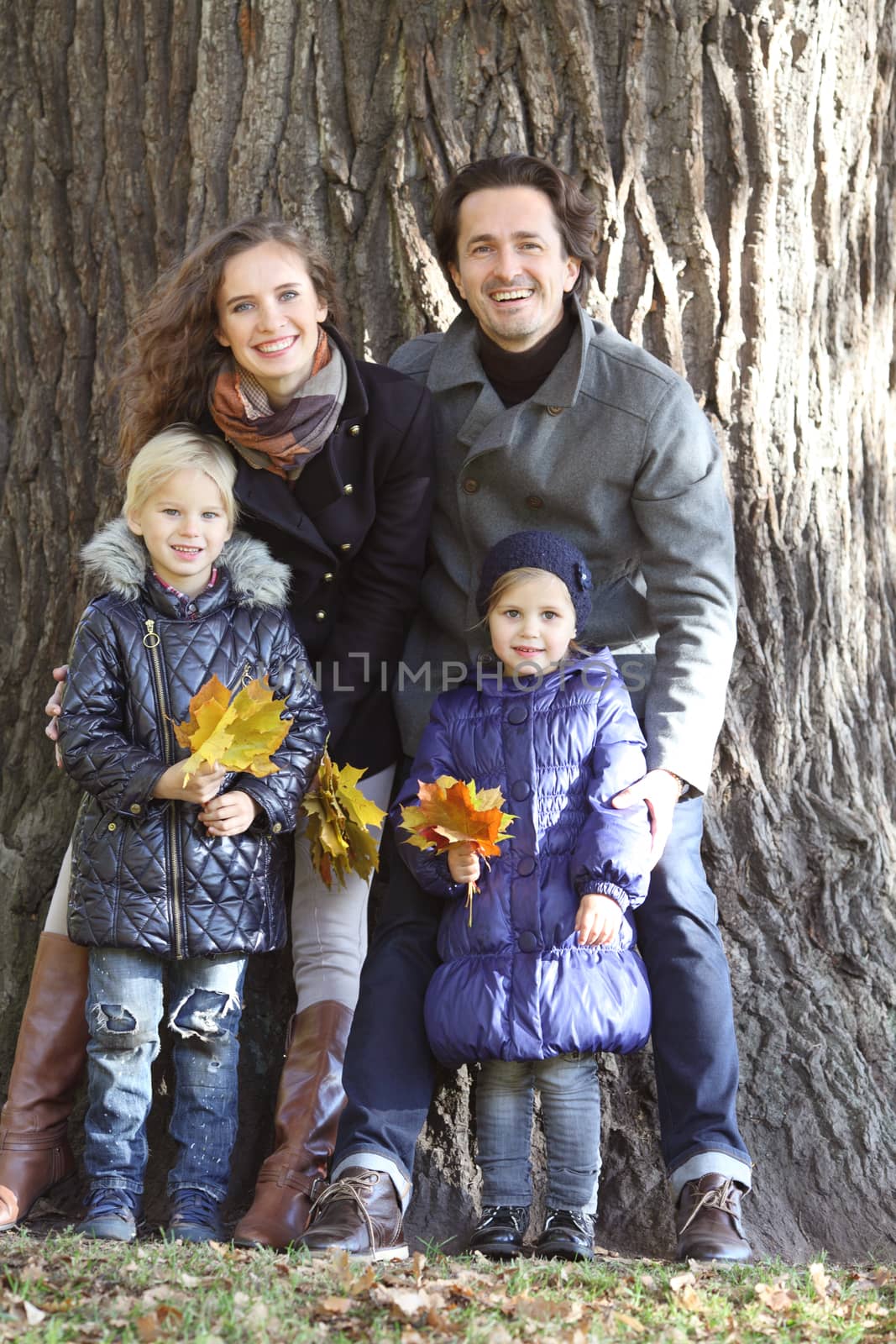 Happy smiling family of parents and children in autumn park standing near big tree trunk