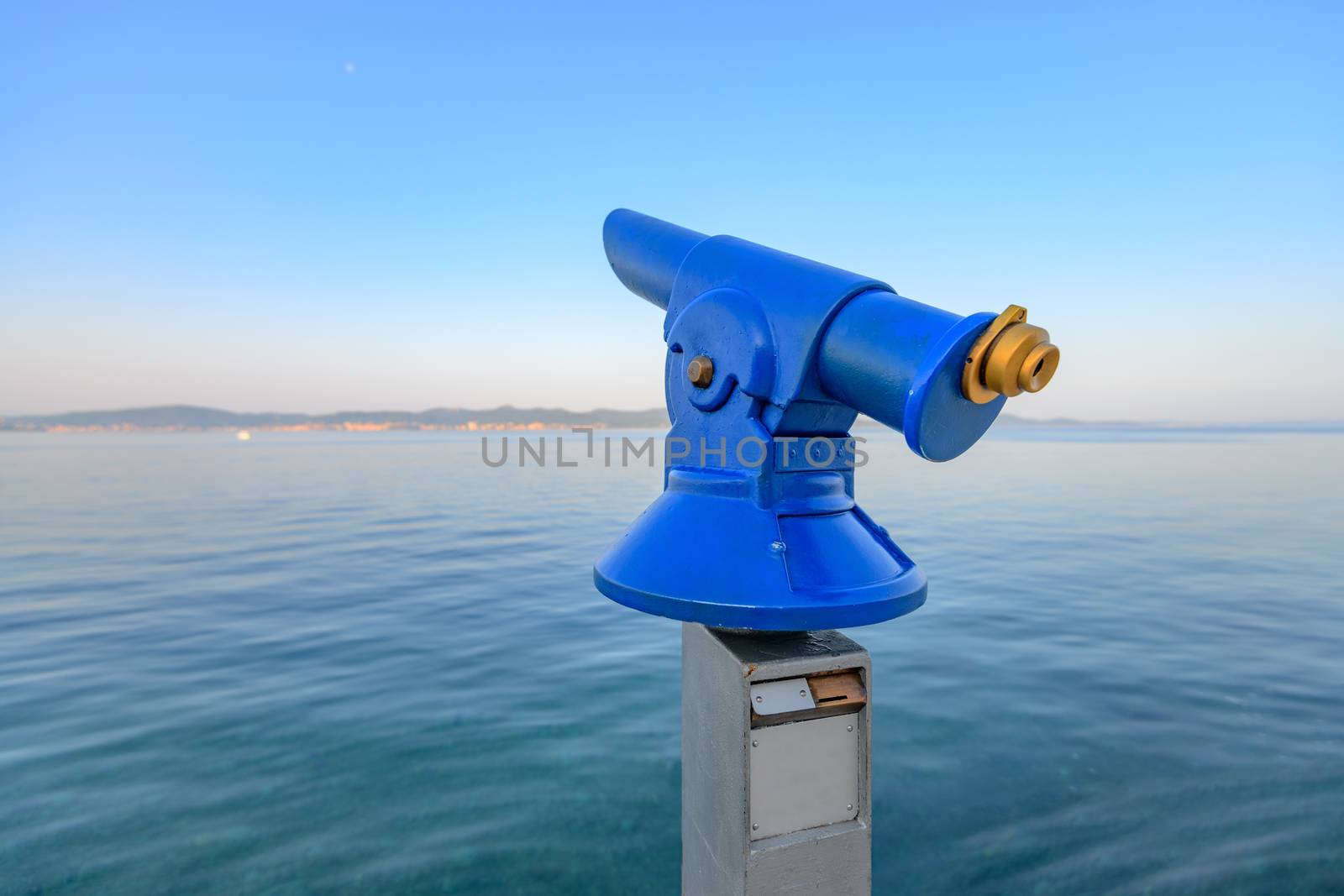 Blue public coin operated telescope, sea and island in background, located on waterfron early in the morning