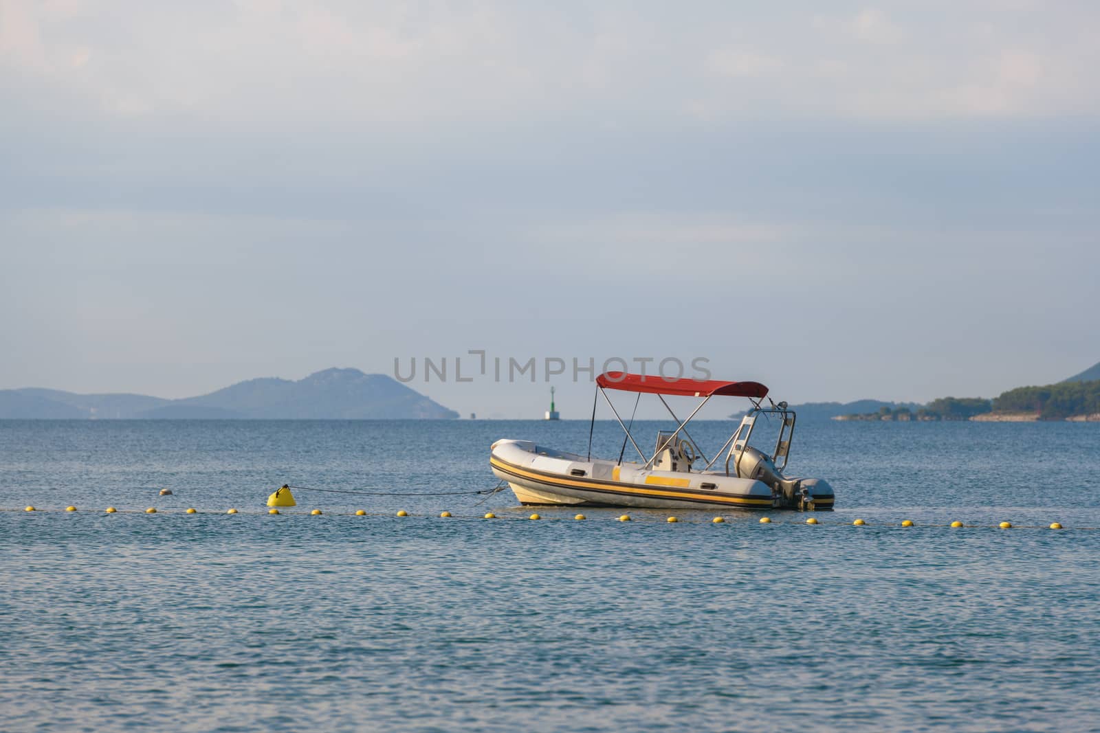 Small rubber motor boat, dinghy in morning light, red sun shade above the boat, moored on buoy, islands in the background