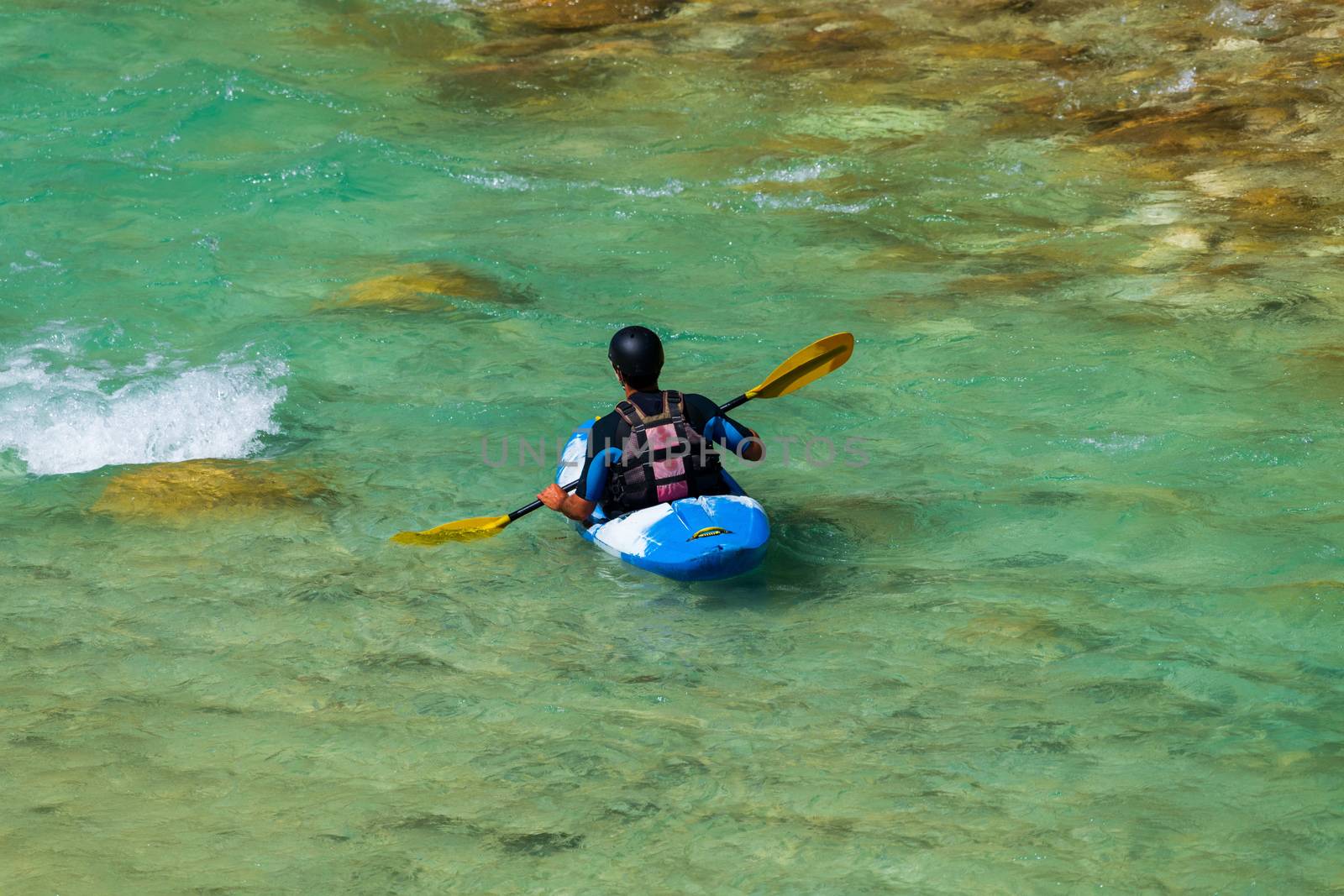 A man kayaking in emerald, turquoise mountain river, wearing wetsuits and avoiding roks and boulders in the streaming water, facing away, blue kayak