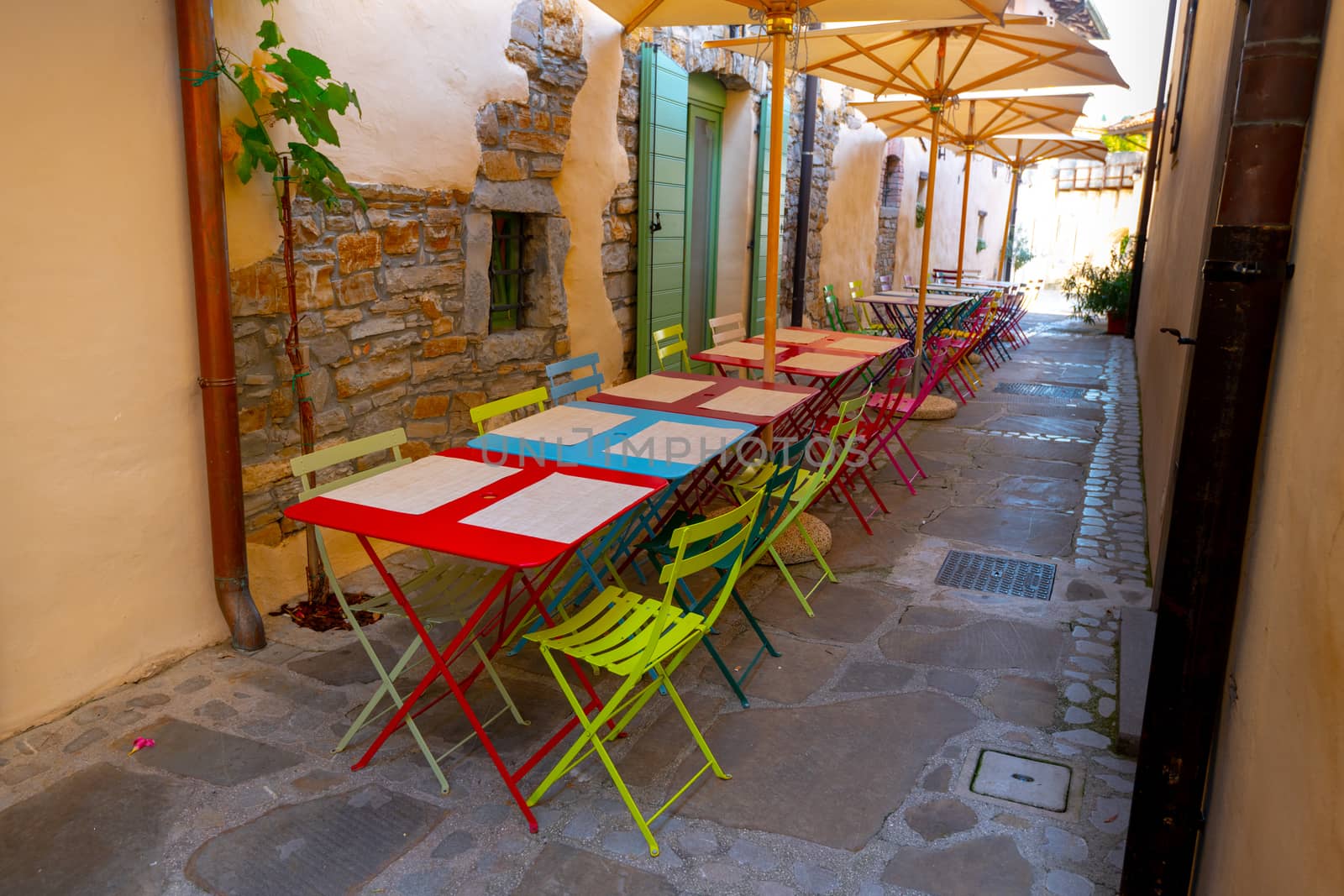 Small caffee, restaurant in narrow street in a medieval town with colorful tables and chairs, nobody, empty, umbrellas for shade