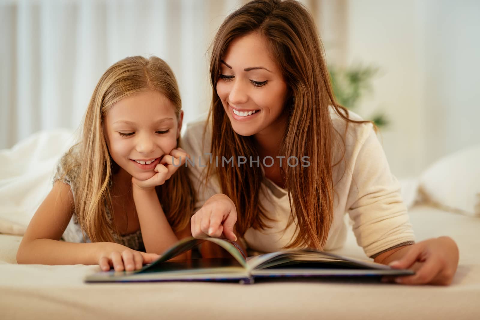 Beautiful smiling mother and her daughter reading book in bed. 