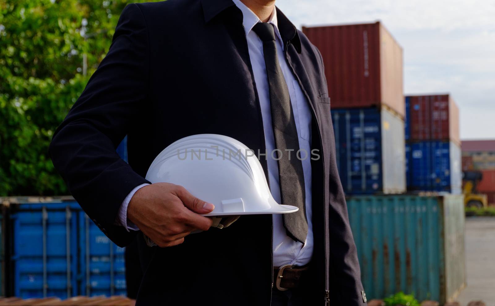 The engineer is holding a helmet on containers background. The construction manager on containers background.