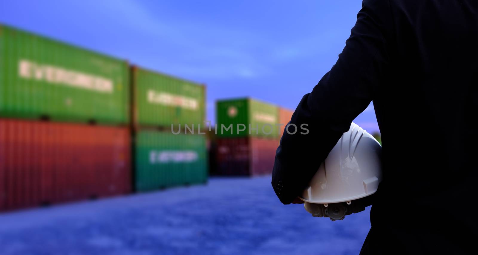 The engineer is holding a helmet on containers background. The construction manager on containers background.