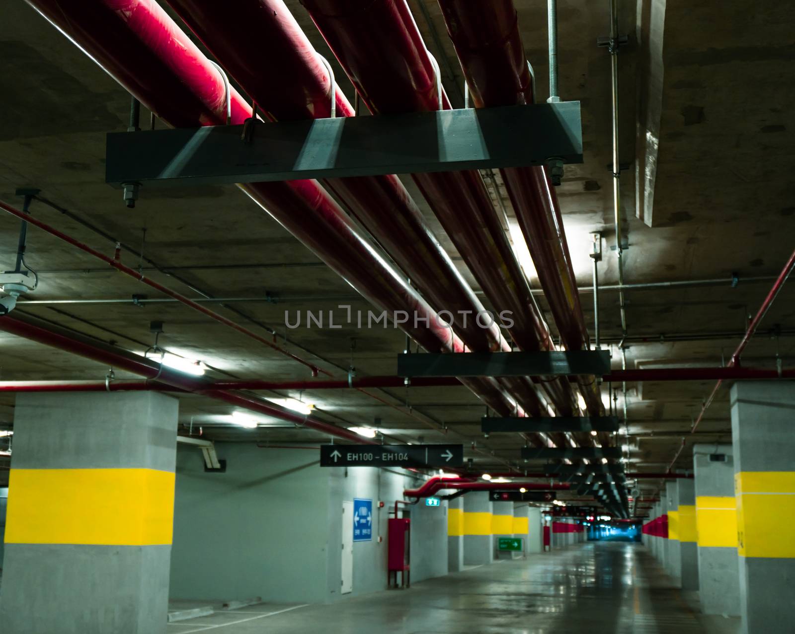Perspective view of empty indoor car parking lot at the mall. Underground concrete parking garage with open lamp at night. Wiring and plumbing in the mall by Fahroni