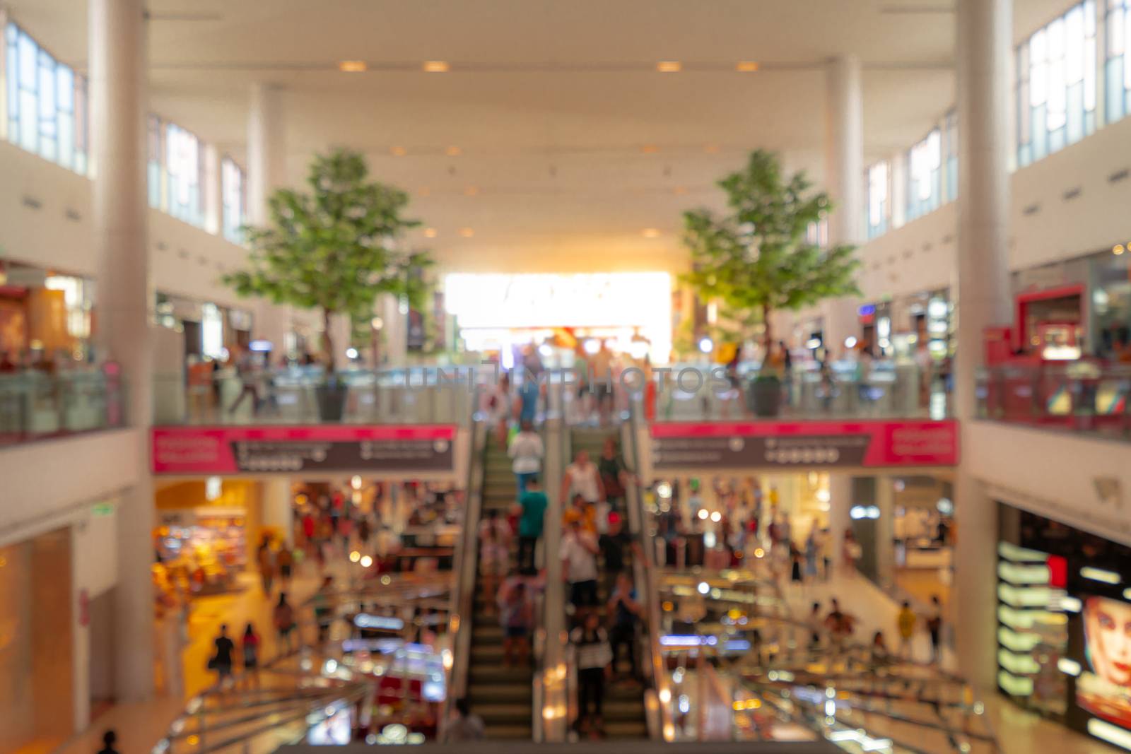 Blurred shopping mall background. People walking and shopping on holiday. People in escalators at the modern shopping mall that second floor decorated with two trees for fresh environment.