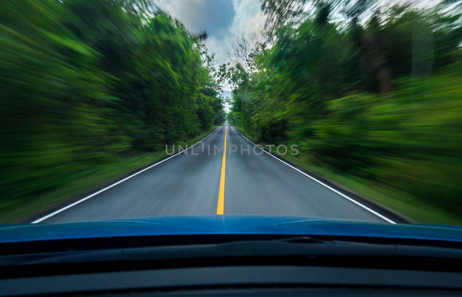 Front view of blue car driving with fast speed on the middle of asphalt road with white and yellow line of traffic symbol in the green forest. Trees beside the road is blurred. Car with blur motion. by Fahroni