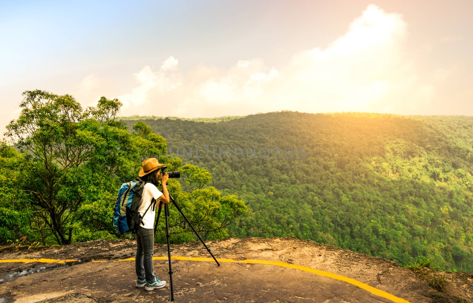 Young traveling woman with backpack hat and camera on tripod stand on the top of the mountain cliff watching beautiful view of woods and sky on her vacation. Asian woman travel alone. by Fahroni
