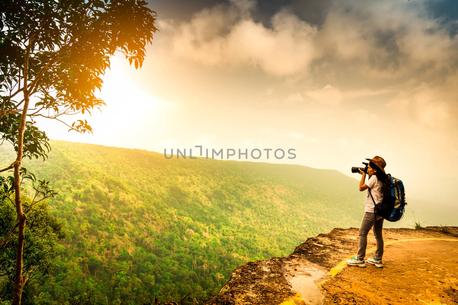 Young traveling woman with backpack, hat and camera stand on the mountain cliff. Female photographer taking picture of tropical forest, sky and clouds on her vacation. Asian woman travel alone. by Fahroni