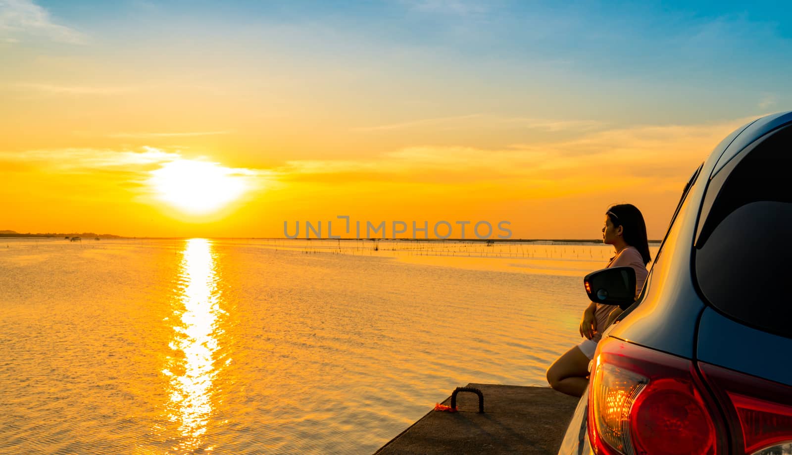 Young Asian woman wear T-shirt, short pants and sunglasses on her head stand leaning blue compact SUV sport car by the sea at sunset with beautiful clouds and yellow-orange sky and reflection on water by Fahroni
